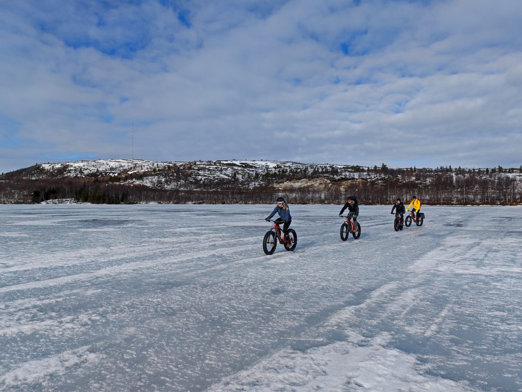 Fire syklister på el-fatbike krysser et islagt vann ved Kirkenes en lys vårvinterdag, med lave åser og skog i bakgrunnen.