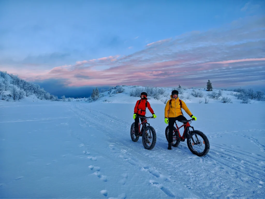 To syklister i røde og gule jakker står med el-fatbiker på en snødekt sti ved Kirkenes. Rosa skyer fyller vinterhimmelen, og det er dyrespor i snøen foran dem.