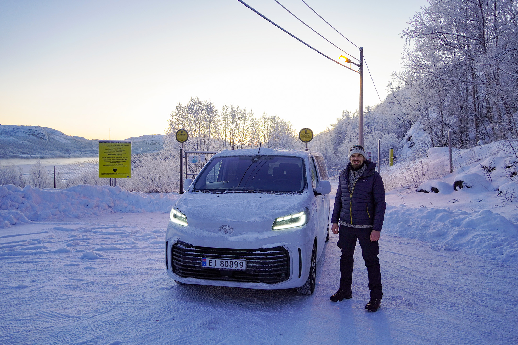Guide standing next to a white minivan at Skafferhullet, a former Cold War border crossing near Kirkenes. Behind them are the gate, yellow warning signs and frost-covered trees on a cold winter morning.