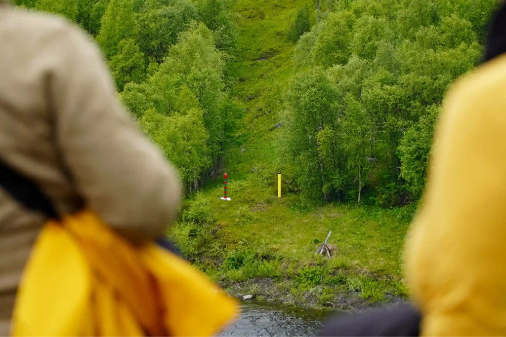 View over the shoulders of two guests towards the border markers across the water near Kirkenes. The Russian and Norwegian border posts stand in the cleared border corridor, which runs up through green birch forest.