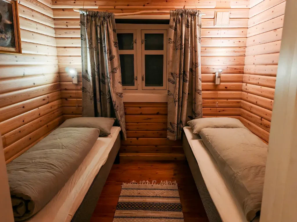 Bedroom with two single beds, log walls, reading lamps, a small window with curtains and a woven rug on the wooden floor at Namdalen Wilderness Lodge in the Pasvik Valley.