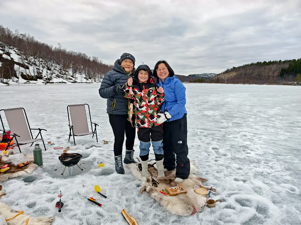 Smiling boy holds up the fish he caught, together with two adults on a reindeer skin on the ice while ice fishing at the Russian border near Kirkenes.