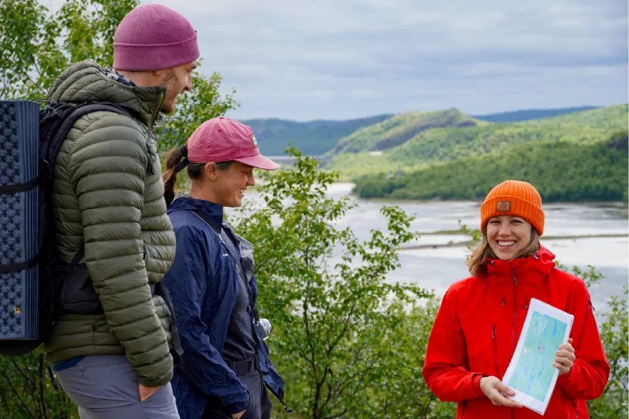 Smiling guide in a red jacket and orange beanie holds a map and tells two guests about the border area, with the Pasvik River and green hills in the background on a summer day near Kirkenes.