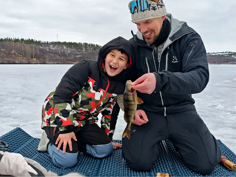 Happy boy laughs as the guide holds up a perch they caught while ice fishing on a border lake near Kirkenes. They kneel on mats on the ice, with snowy hills in the background.