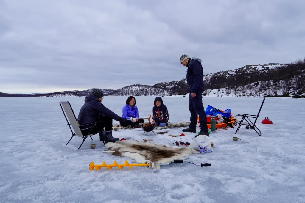 Family and guide gathered around a small campfire on a frozen border lake near Kirkenes, with reindeer skins, chairs, an ice auger and snowy hills around.