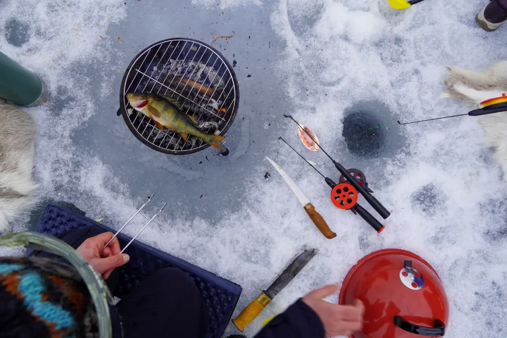 A perch lies on the grill over the fire on the ice, seen from above, with knives, small ice fishing rods and a hole in the ice next to it.