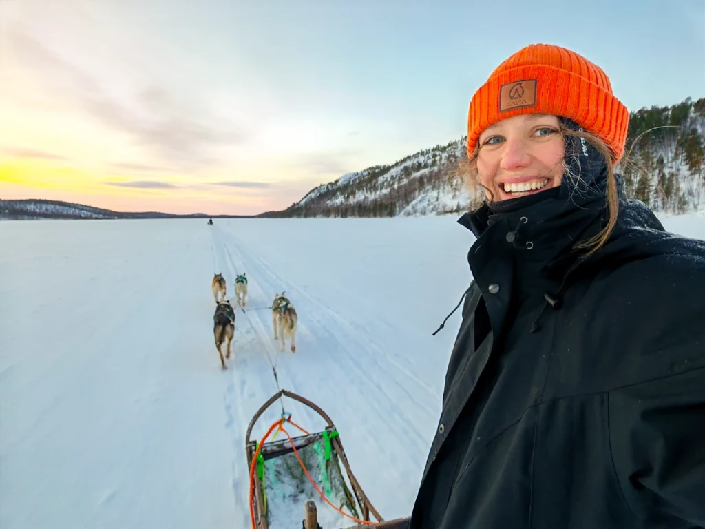 Smiling woman in an orange beanie takes a selfie while driving a dog sled across a frozen lake in the Pasvik Valley, with another team ahead and a golden winter sky over the hills.