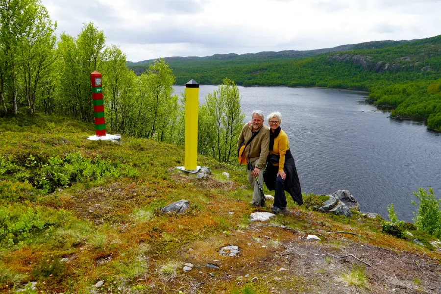 Smiling couple stands by the yellow Norwegian and the red and green Russian border posts on a hike near Kirkenes, with the Pasvik River and green hills in the background.