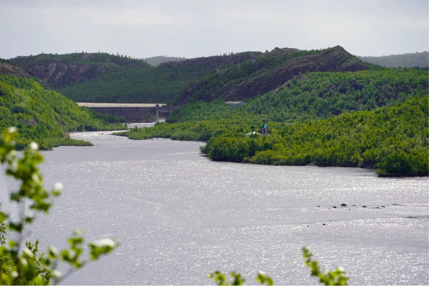View across the Pasvik River to the Boris Gleb Church with its green domes on the Russian side, surrounded by green hills and a hydropower dam in the background on a summer day near Kirkenes.