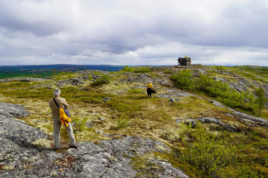 Hikers walk across open, lichen-covered tundra towards a stone cairn on the hilltop in the border area near Kirkenes, with a view over forest and open land under a cloudy summer sky.