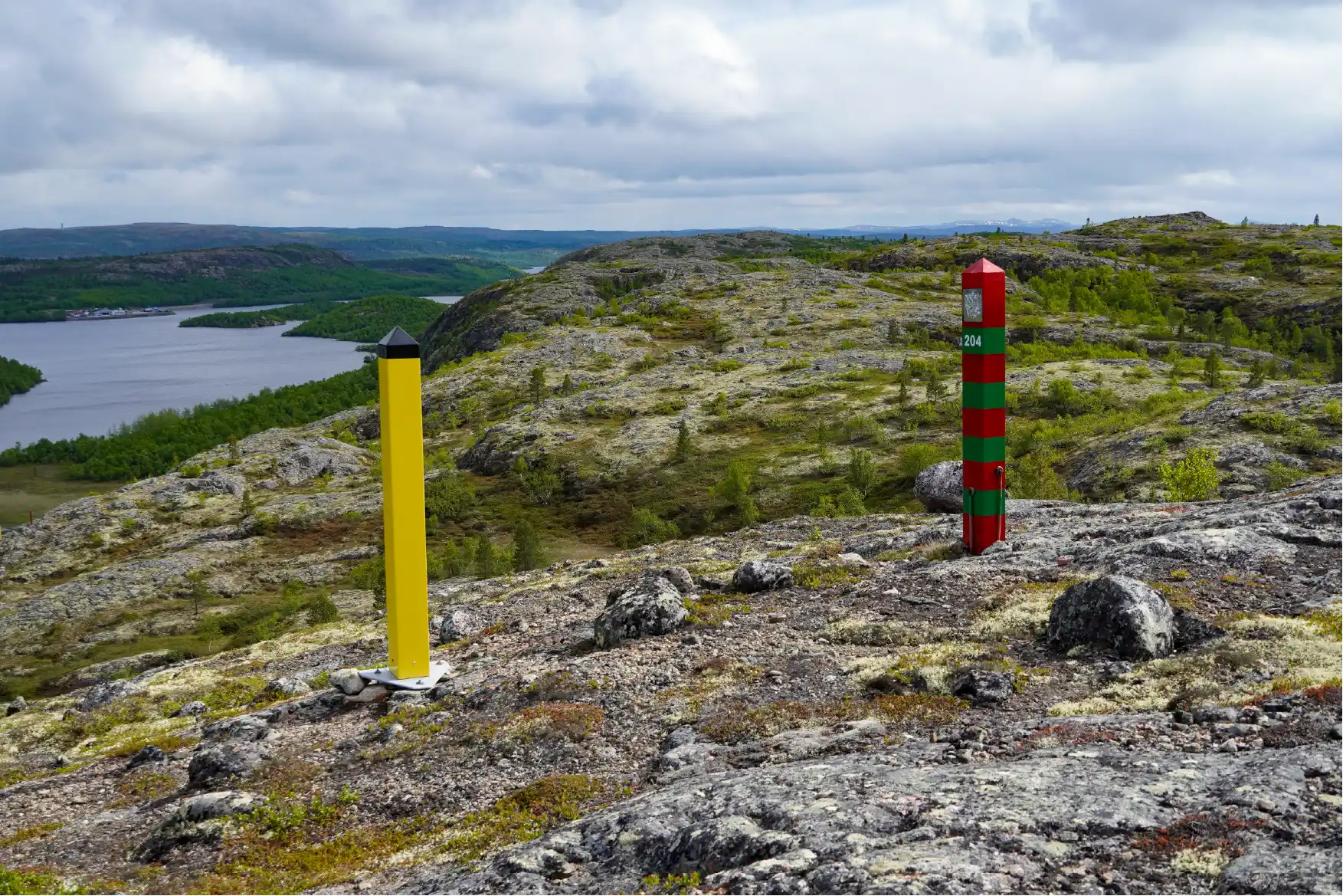 Yellow Norwegian border post and red and green Russian border post with number 204 on lichen-covered rock near Kirkenes on a summer day, with a view over water, forested hills and open tundra.