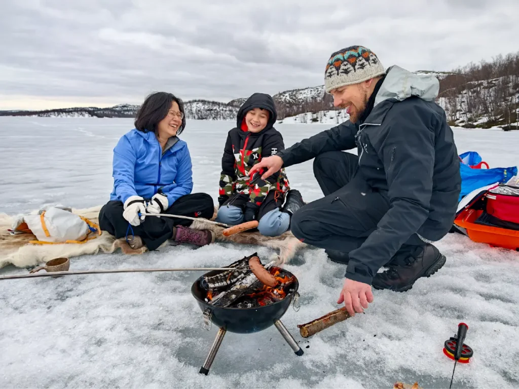 Guide in a knitted hat adds wood to a small campfire on the ice, while a mother and her son sit on reindeer skins grilling reindeer hot dogs during ice fishing at the Russian border near Kirkenes.