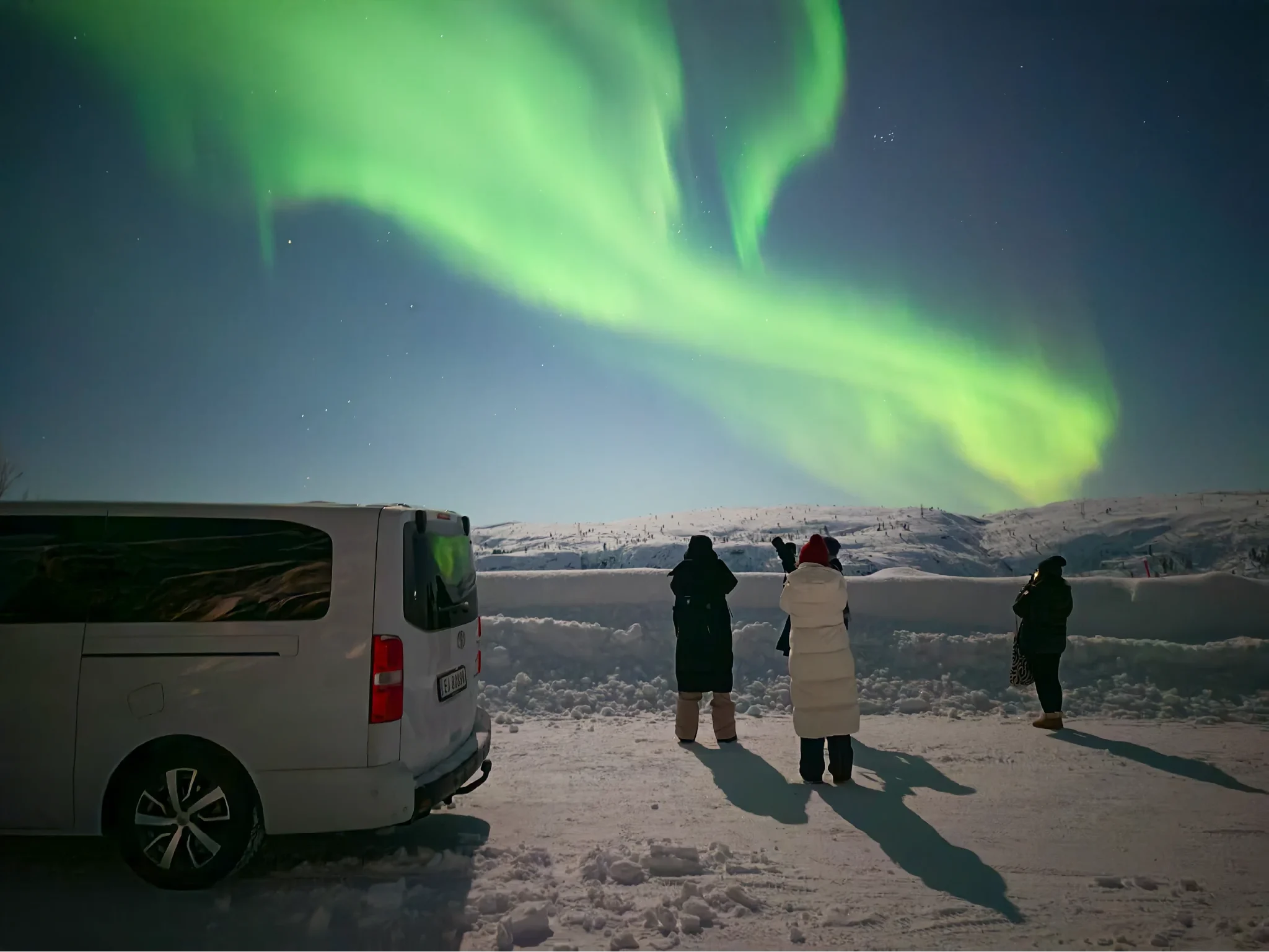 Four guests stand on a snowy road photographing green Northern Lights over white hills near Kirkenes, with a white minivan parked beside them.