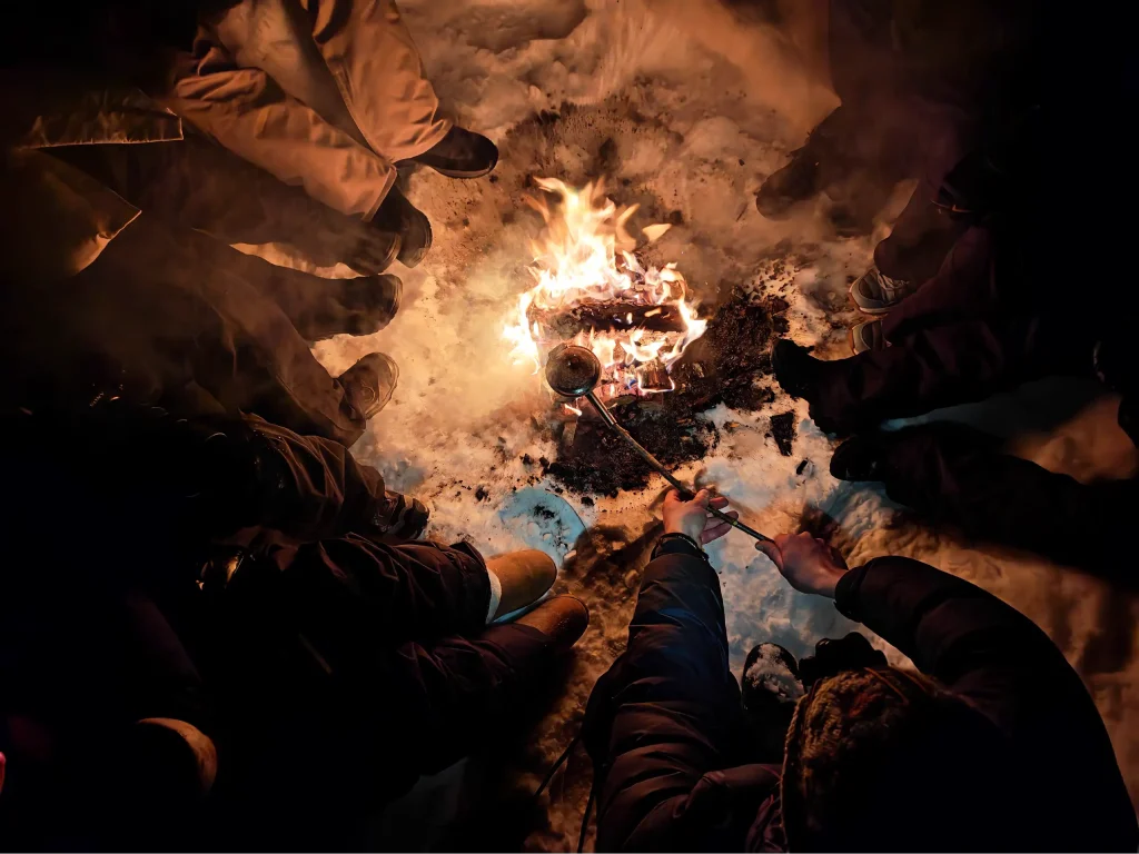 Top-down view of guests sitting around a campfire in the snow, while one holds a long-handled popcorn pan over the flames during a Northern Lights tour near Kirkenes.