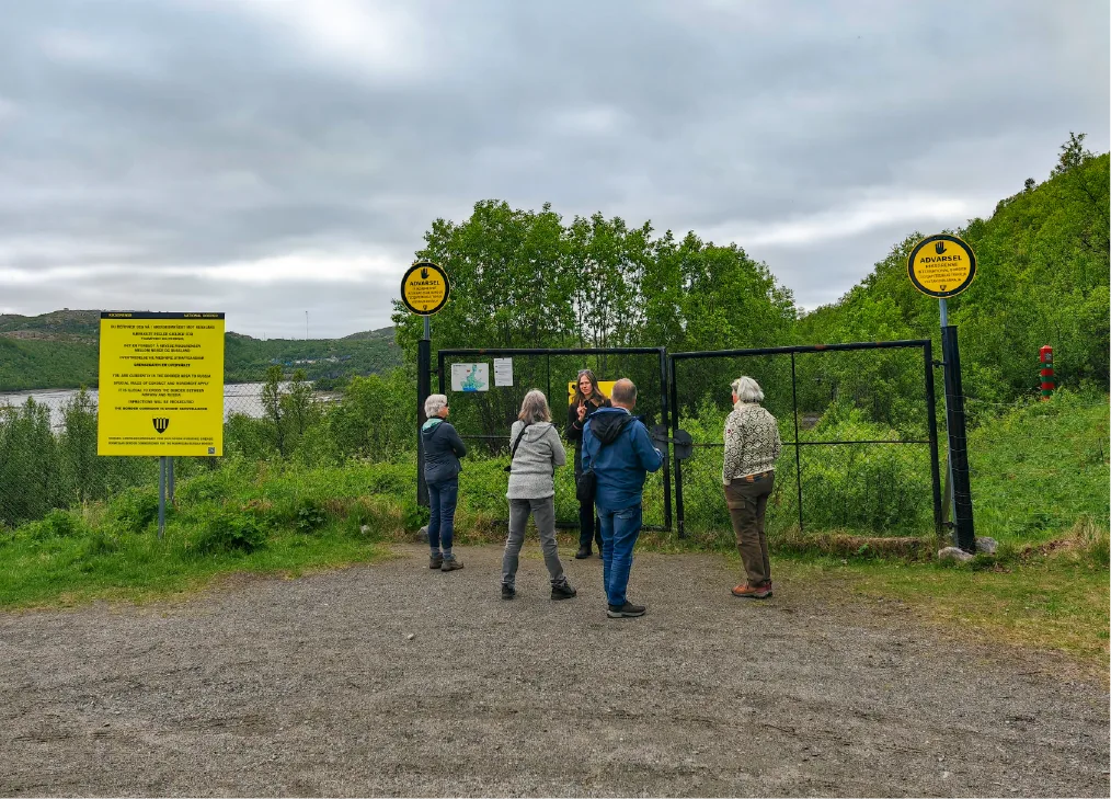 Guide forteller fire gjester om Skafferhullet, en gammel grenseovergang fra den kalde krigen ved russergrensa nær Kirkenes, med stengt port og gule varselskilt.