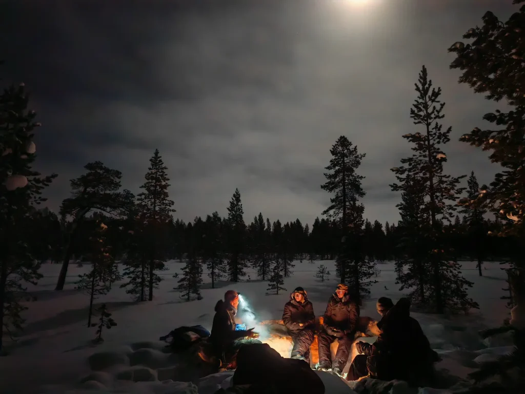 Four people in snowsuits and headlamps sit around a campfire in the snow in a pine forest in the Pasvik Valley, under a cloudy sky with a full moon.